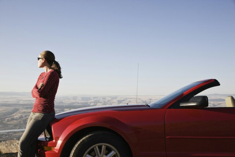 A young Caucasian woman leaning on her convertible sports car at a rest stop.